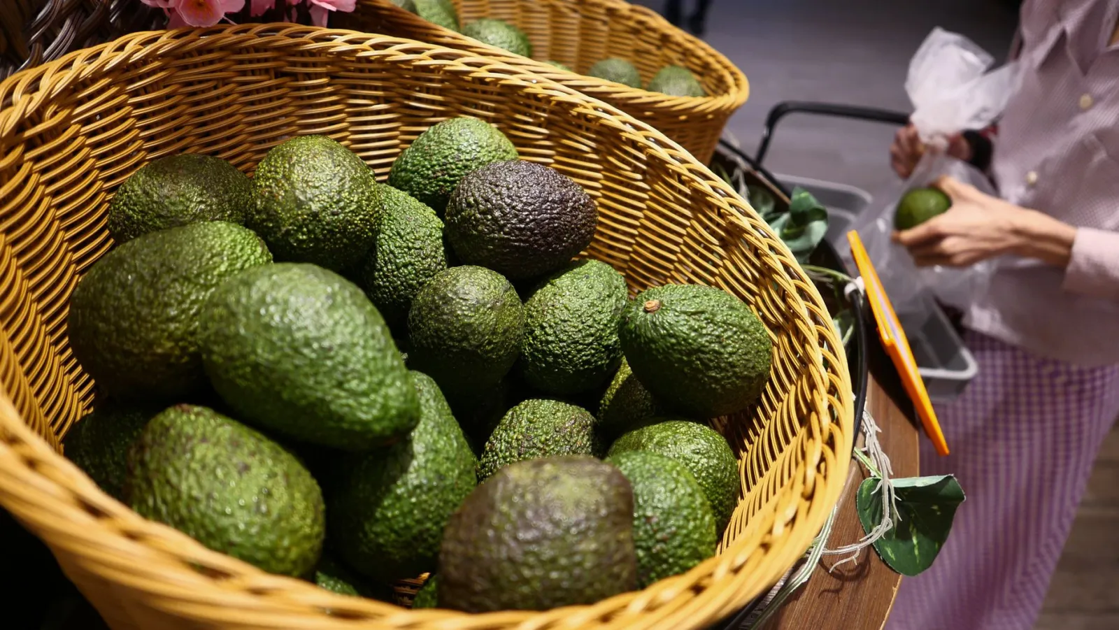 Close-up of fresh avocados in wicker baskets at a market with hands selecting produce.